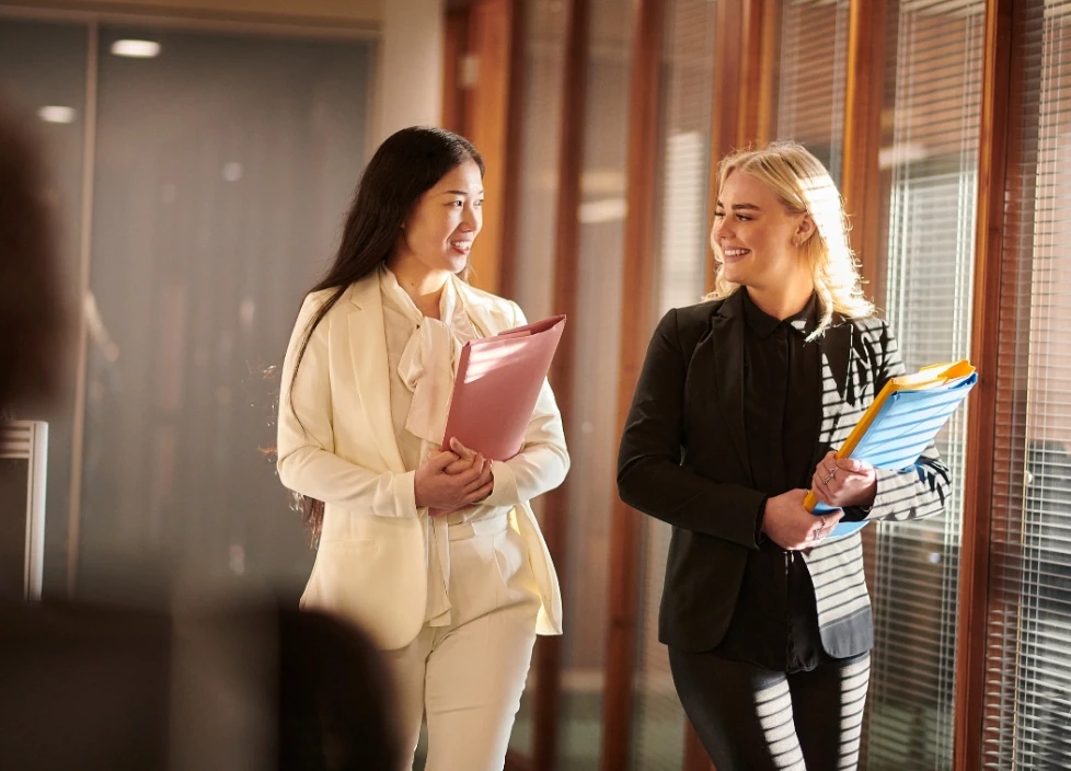 Two female teachers walking through a school hallway while talking happily and holding paper folders