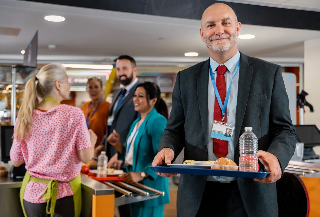 Smiling headmaster in a school canteen, carrying a lunch tray. Teaching staff are in the background with their lunches