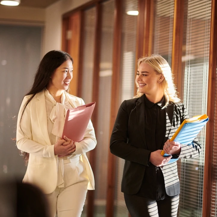 Two women walking through a school hall, carrying paper folders